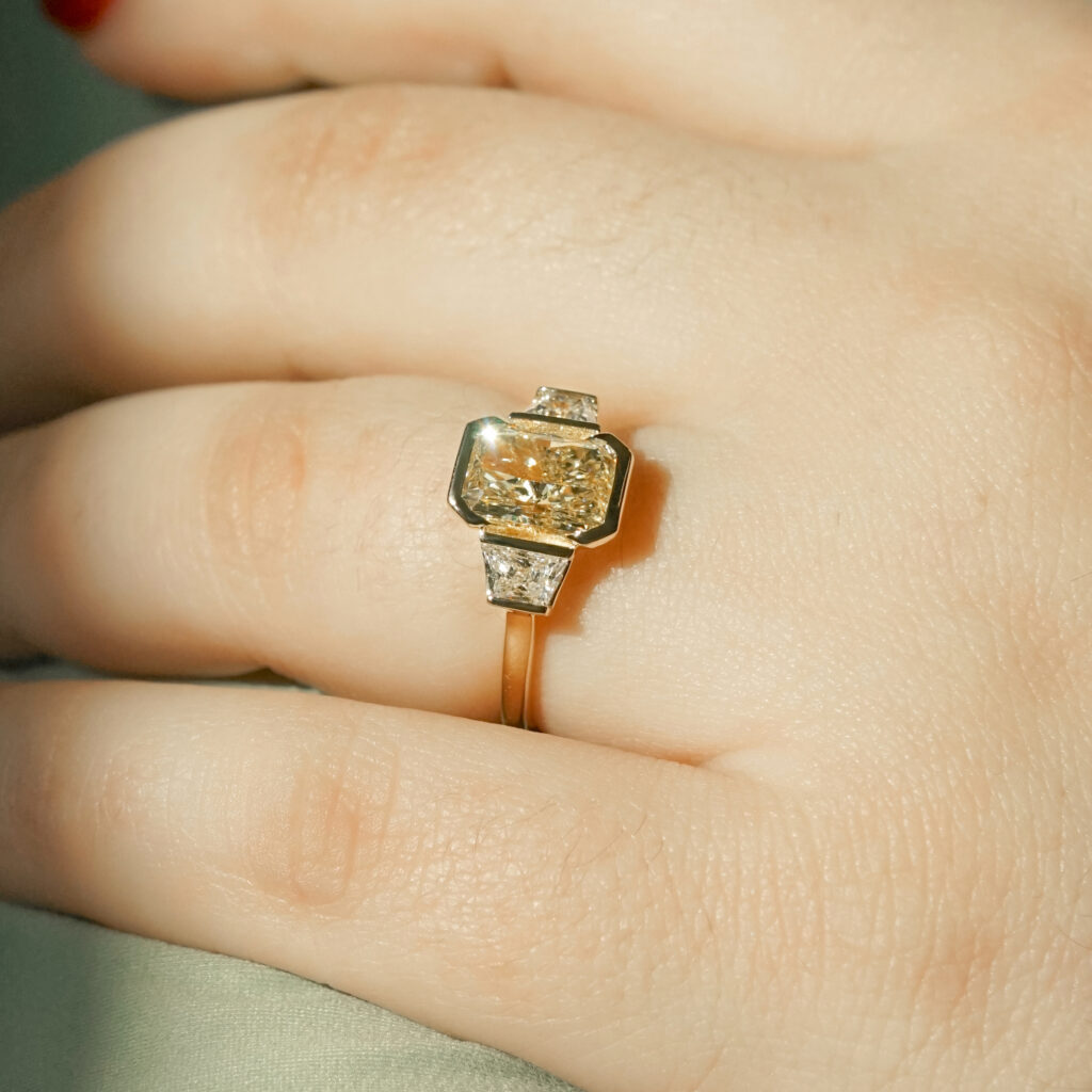 A champagne radiant cut, bezel set diamond with two trapezoid side stones
