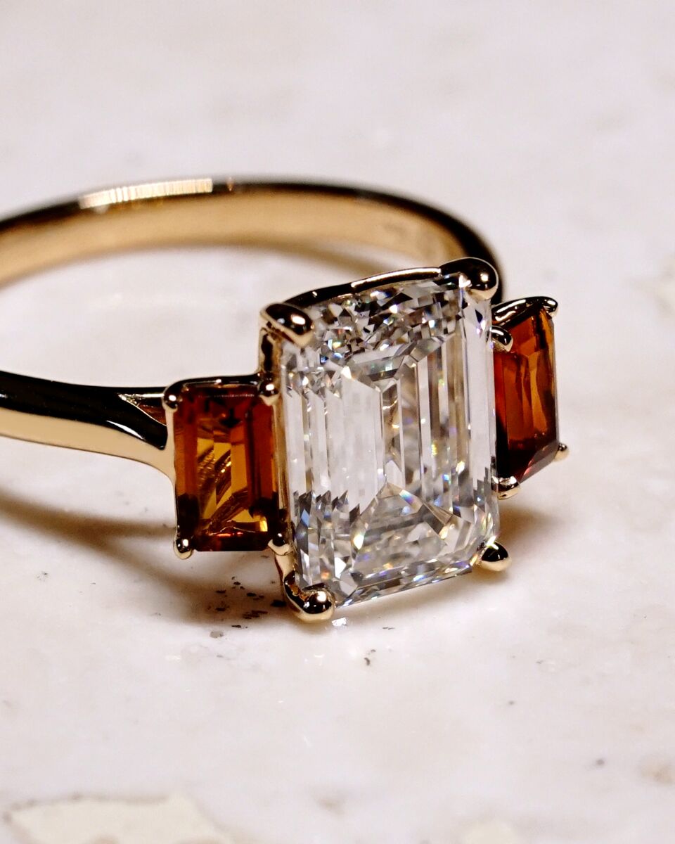 A three-stone emerald cut diamond engagement ring. On either side of the center emerald diamond are two cognac gemstones. 