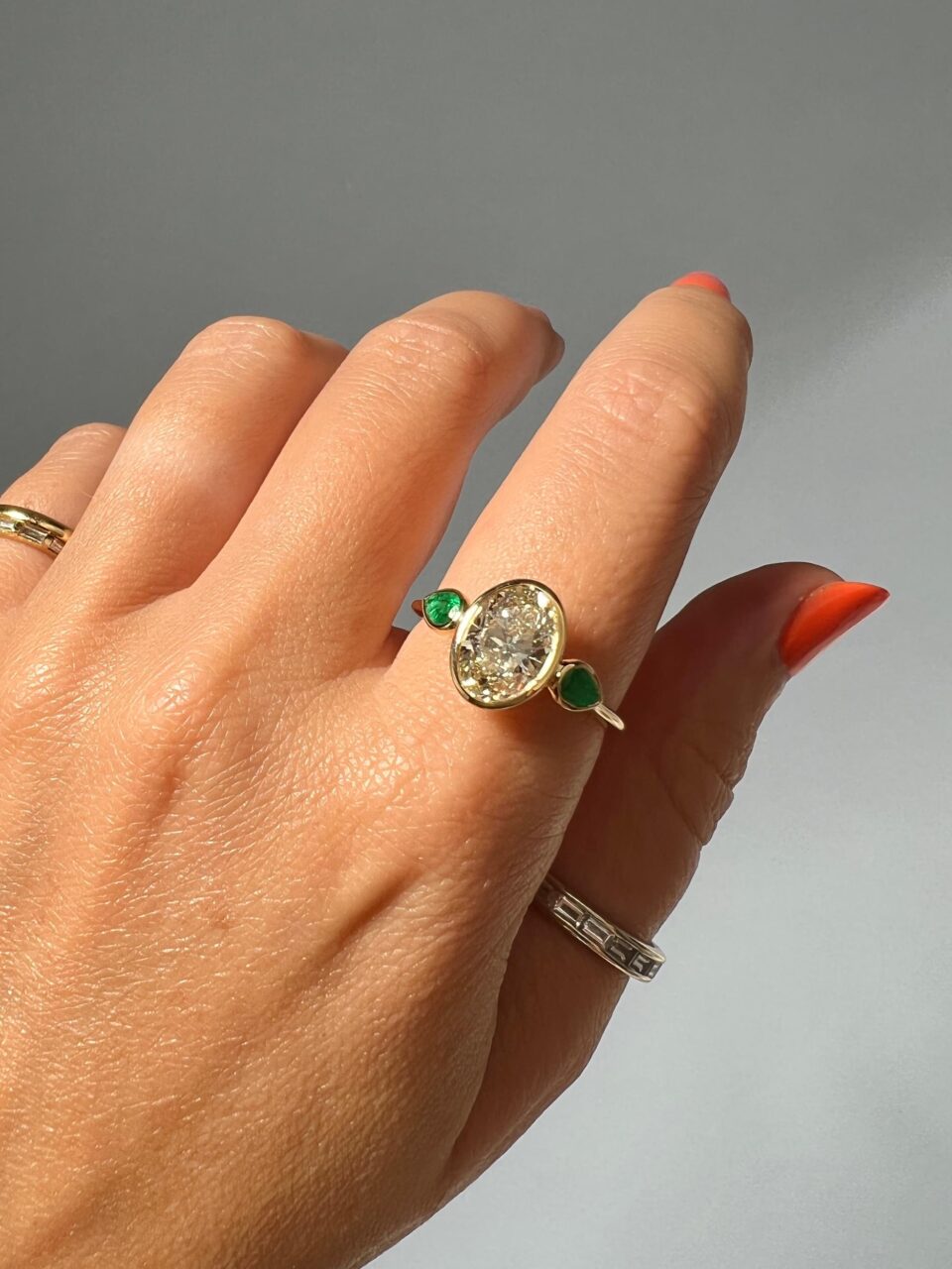 A bezel set oval shaped diamond is framed by 2 pear cut emeralds 