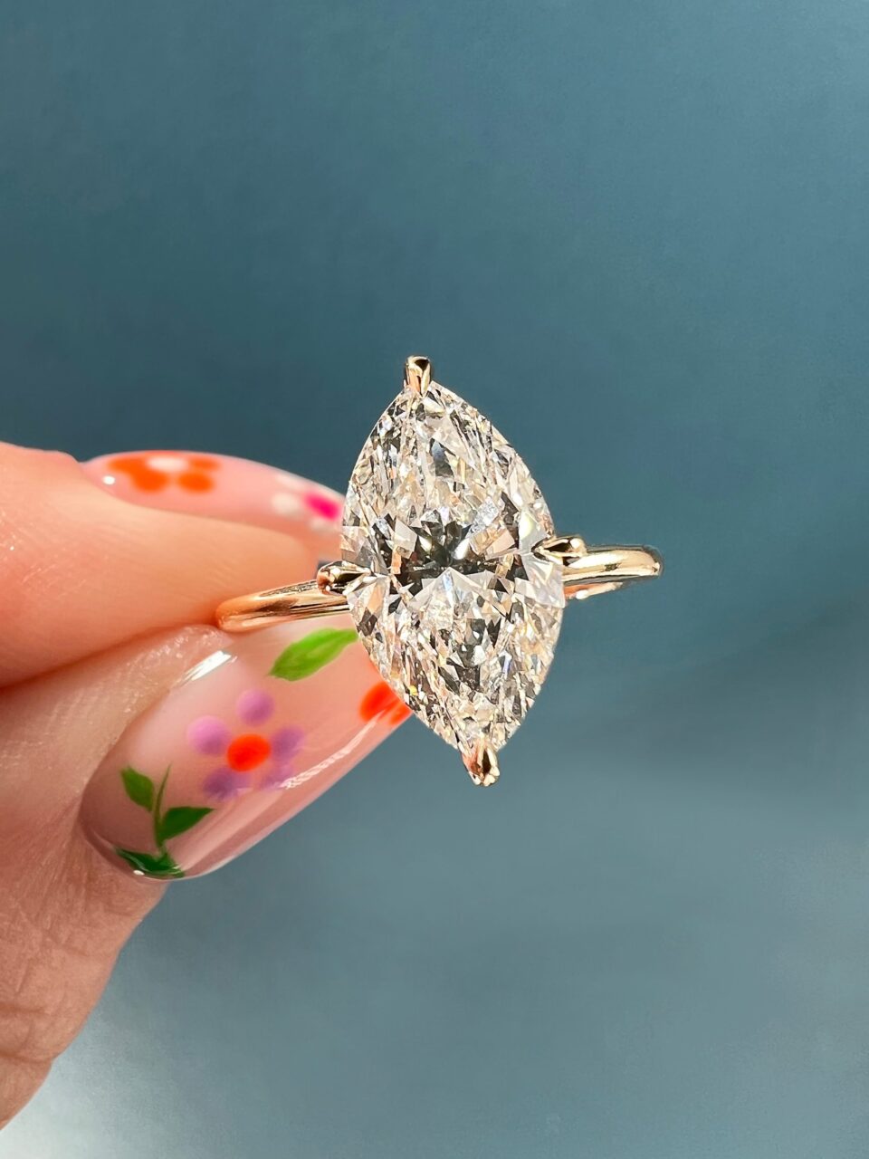 A solitaire Marquise diamond engagement ring is held delicately between two fingers decorated with flower printed nails. 